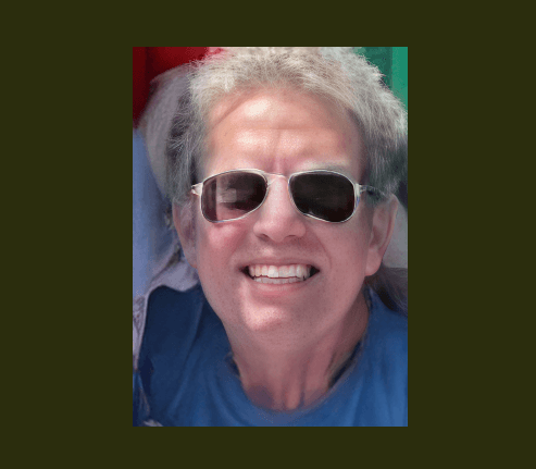 Close-up portrait of a smiling person with light hair wearing dark sunglasses and blue shirt.